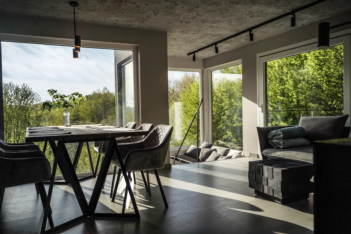 A bright and airy dining area features a spacious wooden table surrounded by stylish chairs. Large windows provide a panoramic view of the lush greenery outside, allowing natural light to illuminate the room. A cozy seating area with soft cushions is positioned nearby.