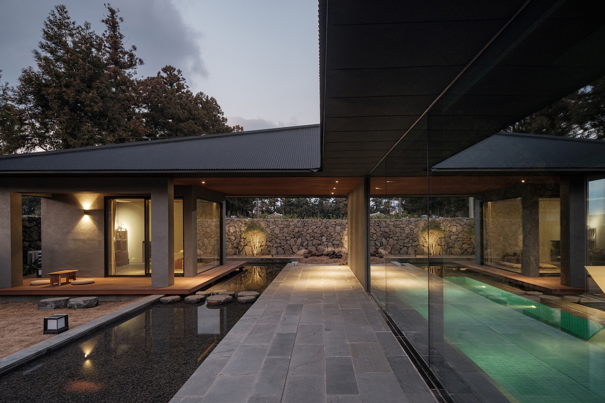 An outdoor view showcases a tranquil space featuring a glass-walled structure connected to a warm-toned wooden roof. Reflective surfaces of the water and swimming pool are visible, alongside natural stone pathways and serene landscaping, inviting guests to enjoy the peaceful surroundings.