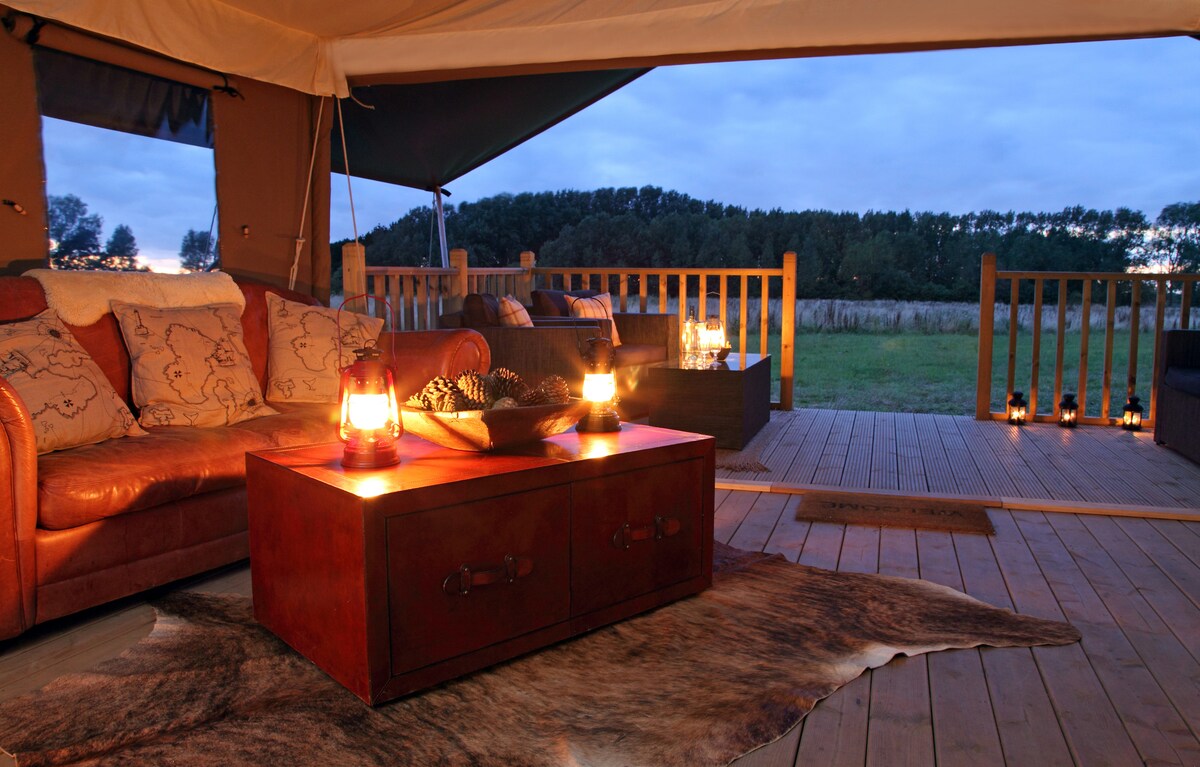 A cozy seating area is featured under canvas, illuminated by soft lantern light. A wooden coffee table rests on a patterned rug, while outdoor seating is visible through the open entrance. Surrounding natural views offer a serene backdrop to the inviting interior.