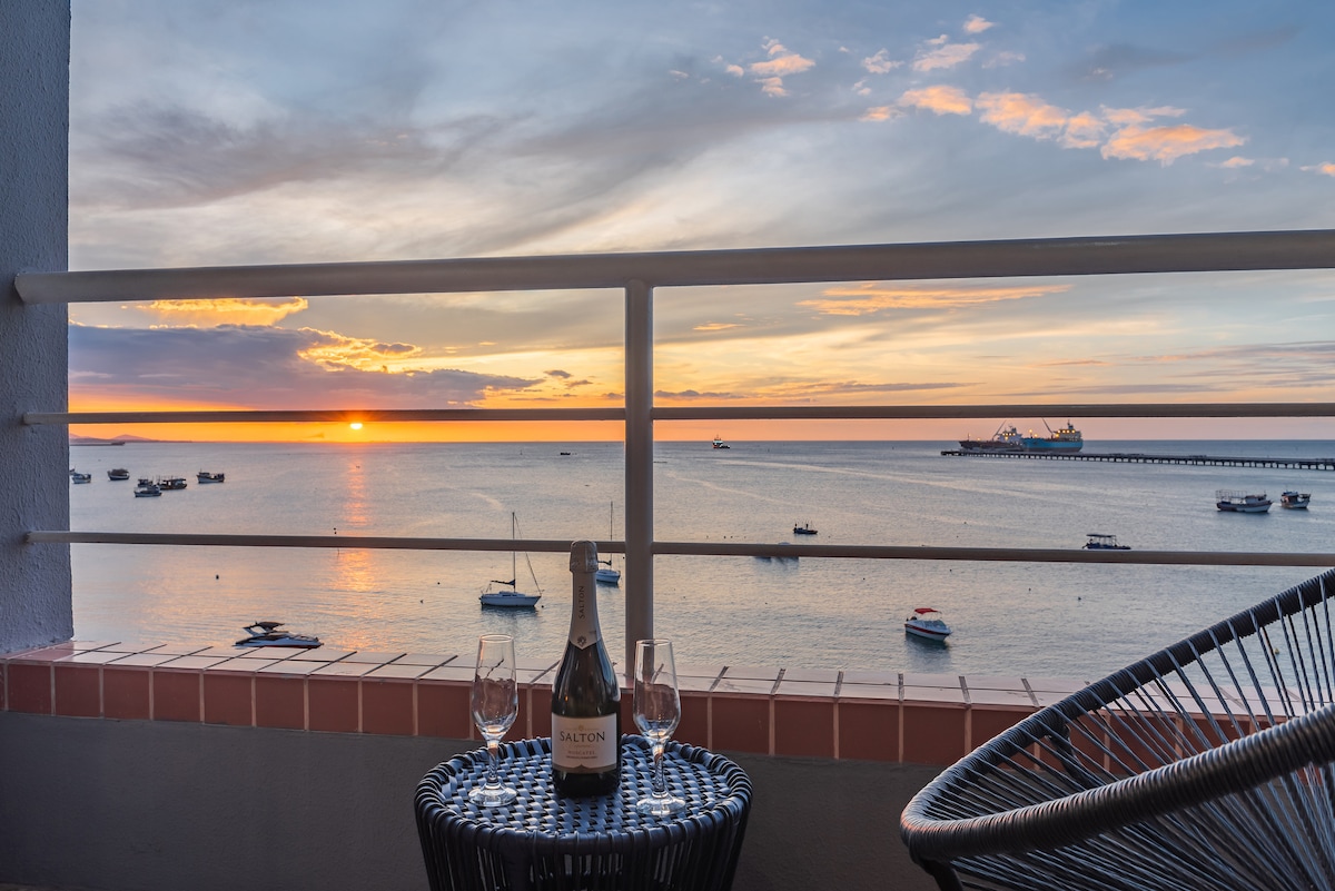 A balcony setting features a bottle of sparkling wine and two glasses on a round black table. The view showcases a vast ocean reflecting hues of orange and purple from the sunset, with boats gently floating in the water and a distant pier visible.