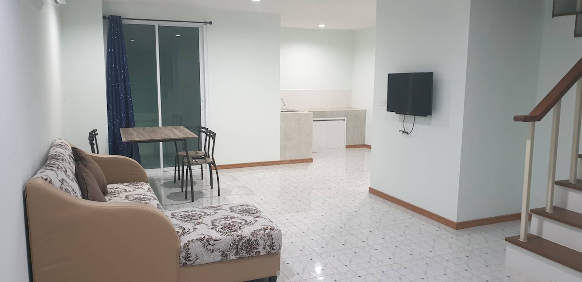 An open-plan living area is shown, featuring a light-colored sofa with patterned upholstery. A dining table with four black chairs is set nearby. A kitchen area is visible in the background, with simple cabinetry and appliances. Large glass doors lead to the outdoor space.
