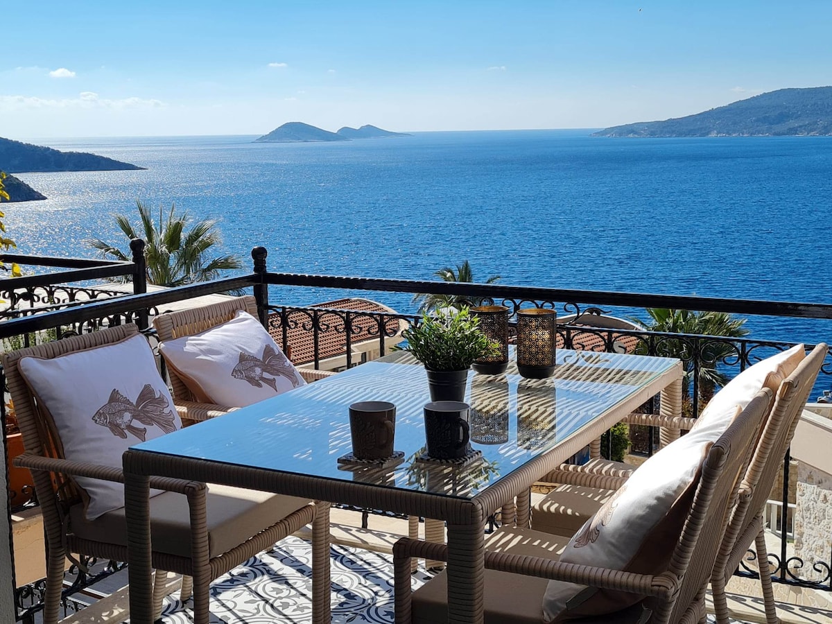 A spacious balcony features a glass-top dining table surrounded by comfortable wicker chairs. Two coffee cups sit on the table, and a planter with greenery adds a natural touch. The panoramic view shows tranquil blue waters with distant islands under a clear sky.