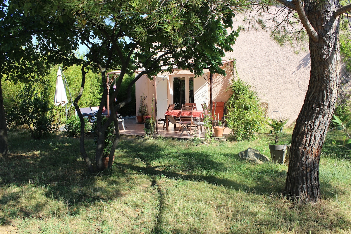 A serene outdoor space is seen, featuring a terrace with wooden loungers and a table. Surrounding plants and trees create a natural setting, while a parasol provides shade. The house is partially visible, offering a glimpse of the welcoming design.
