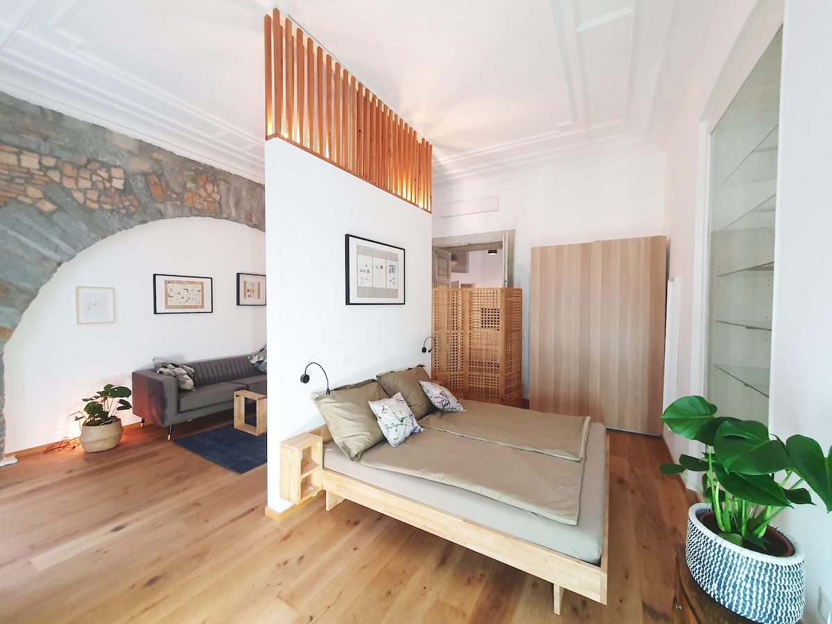 A seating area with a comfortable sofa is paired with a wooden bed featuring neutral bedding and decorative pillows. An archway reveals a stone accent wall, while wood slats provide a touch of separation. Houseplants offer a hint of greenery, complementing the light wooden flooring.