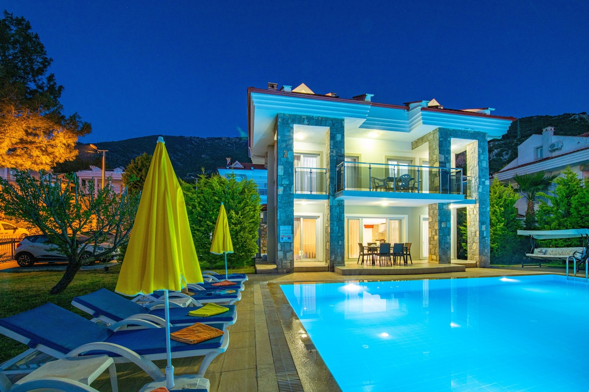 The villa is illuminated at night, showcasing a large pool surrounded by sun loungers and umbrellas. The two-story structure features a balcony and outdoor dining area, with landscaped greenery and trees enhancing the setting.