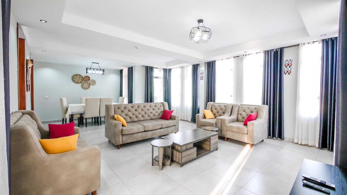 A spacious living area is furnished with two light-colored sofas, each adorned with colorful accent pillows. A low coffee table sits in the center, and large windows allow natural light to brighten the space, which features modern decor and a dining area in the background.
