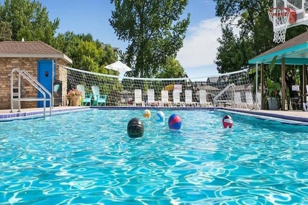 A vibrant pool area is shown, featuring clear turquoise water and several colorful beach balls floating on the surface. Surrounding the pool are lounge chairs and a volleyball net, with greenery and a clear blue sky adding to the refreshing ambiance.