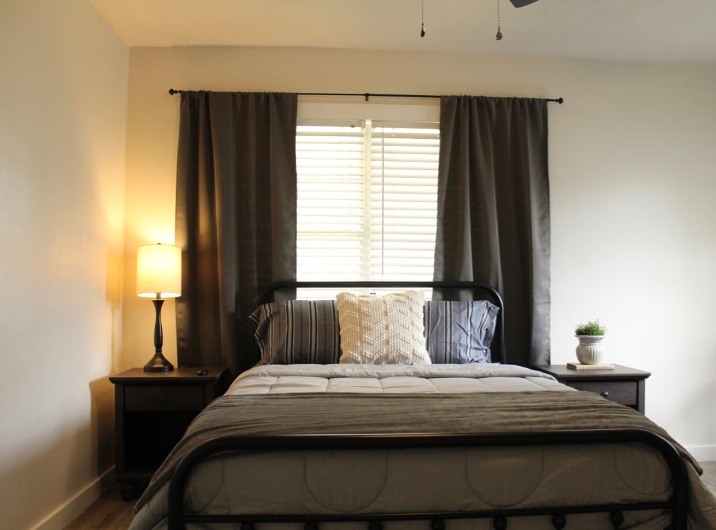 A serene bedroom features a spacious bed adorned with neutral bedding and decorative pillows. Soft light emanates from a bedside lamp, while dark curtains frame a window fitted with light-colored slats. A small potted plant adds a touch of greenery on the nightstand.