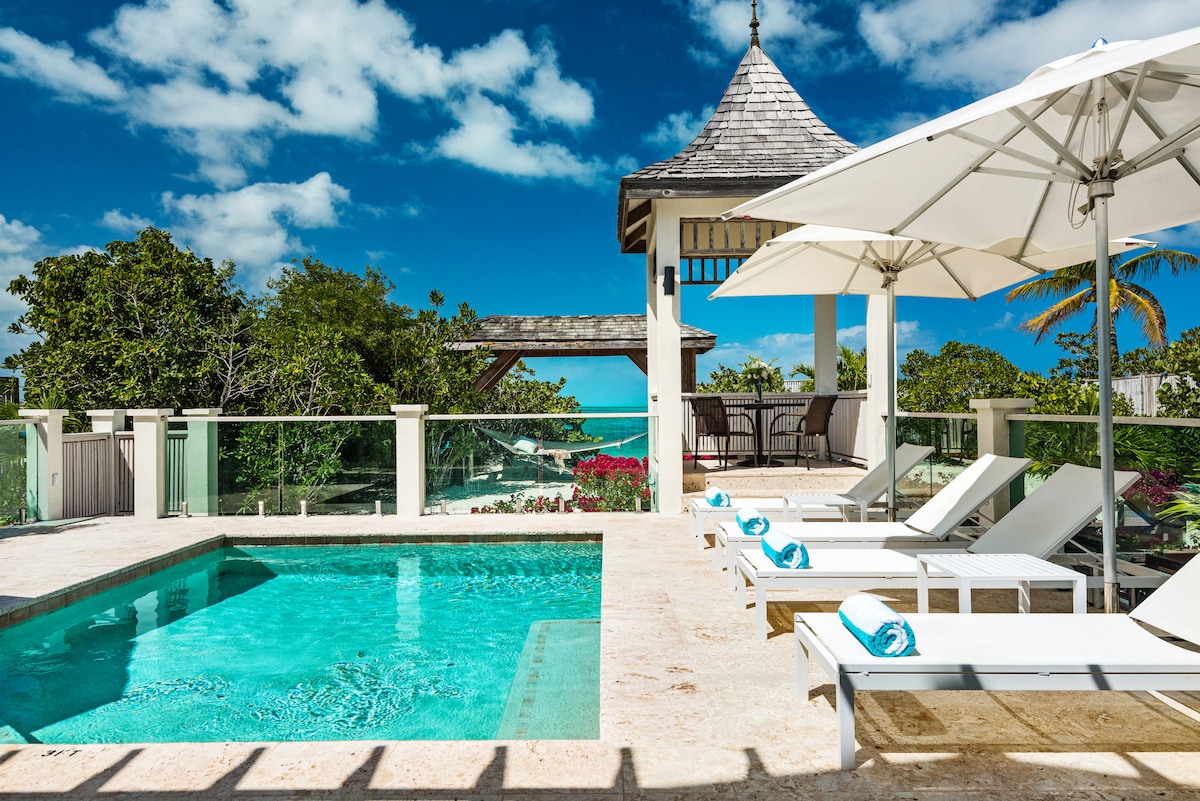 A clear blue pool is surrounded by sun loungers feature plush towels. A gazebo offers shaded seating, complementing the picturesque outdoor area. Lush greenery is visible in the background, with an expansive sky filled with soft clouds above.