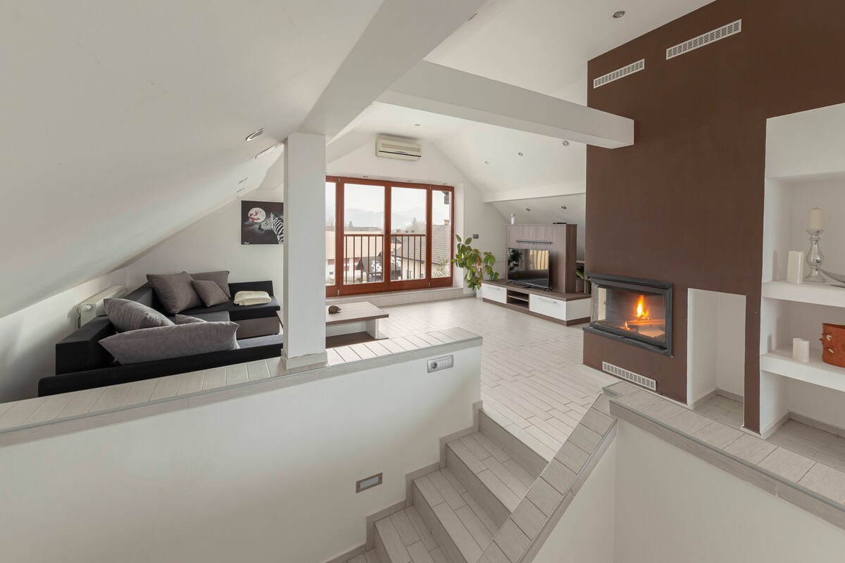 An open and spacious living room is highlighted by a cozy fireplace and large windows that offer natural light. A comfortable dark sofa is positioned nearby, complemented by a decorative wall and minimalist shelving units. Soft gray tones enhance the modern design.