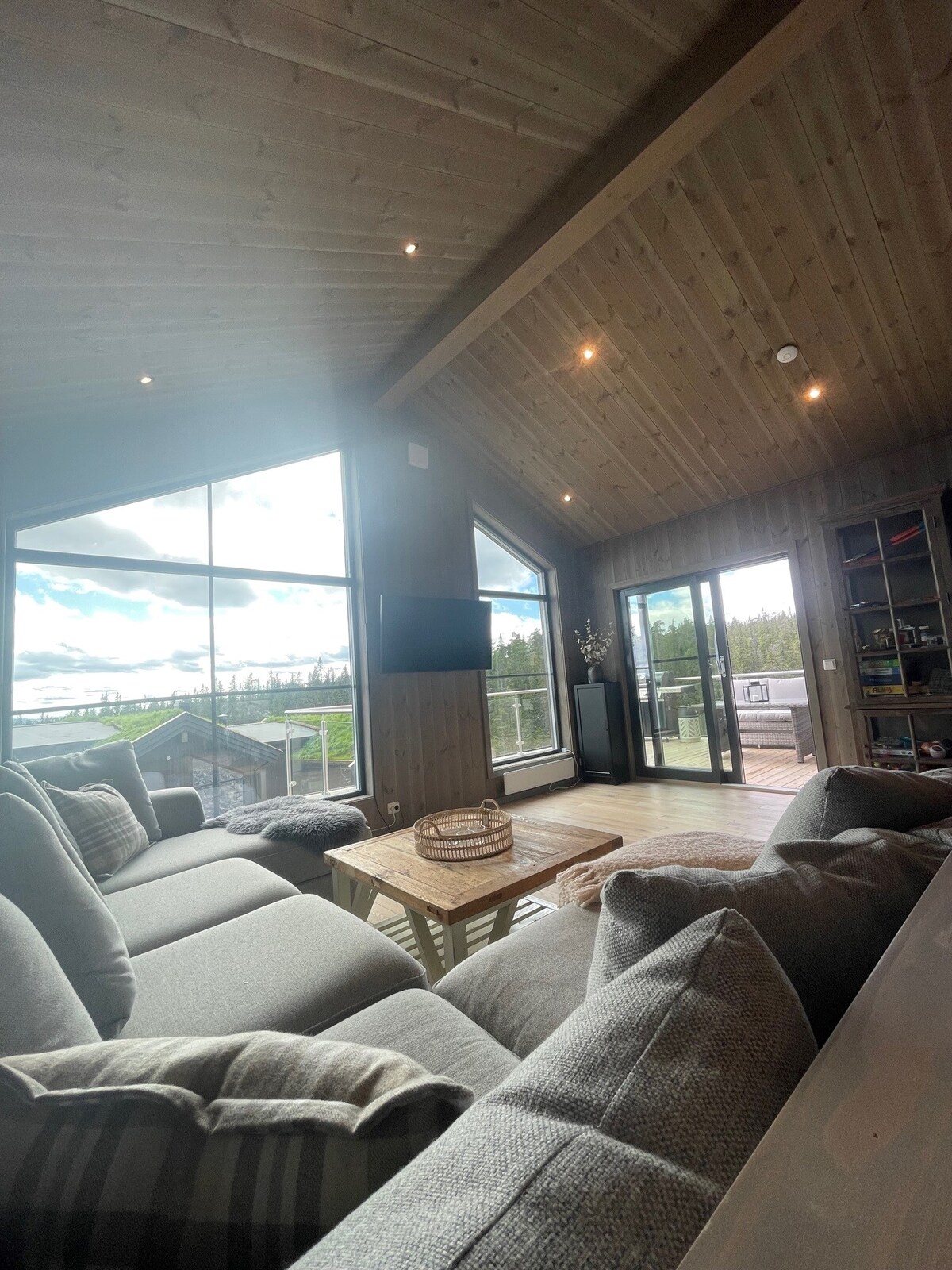 A modern living area features large panorama windows that offer views of the landscape. A low wooden coffee table sits at the center, surrounded by comfortable grey sofas. The ceiling has a natural wood finish, with recessed lighting enhancing the space.