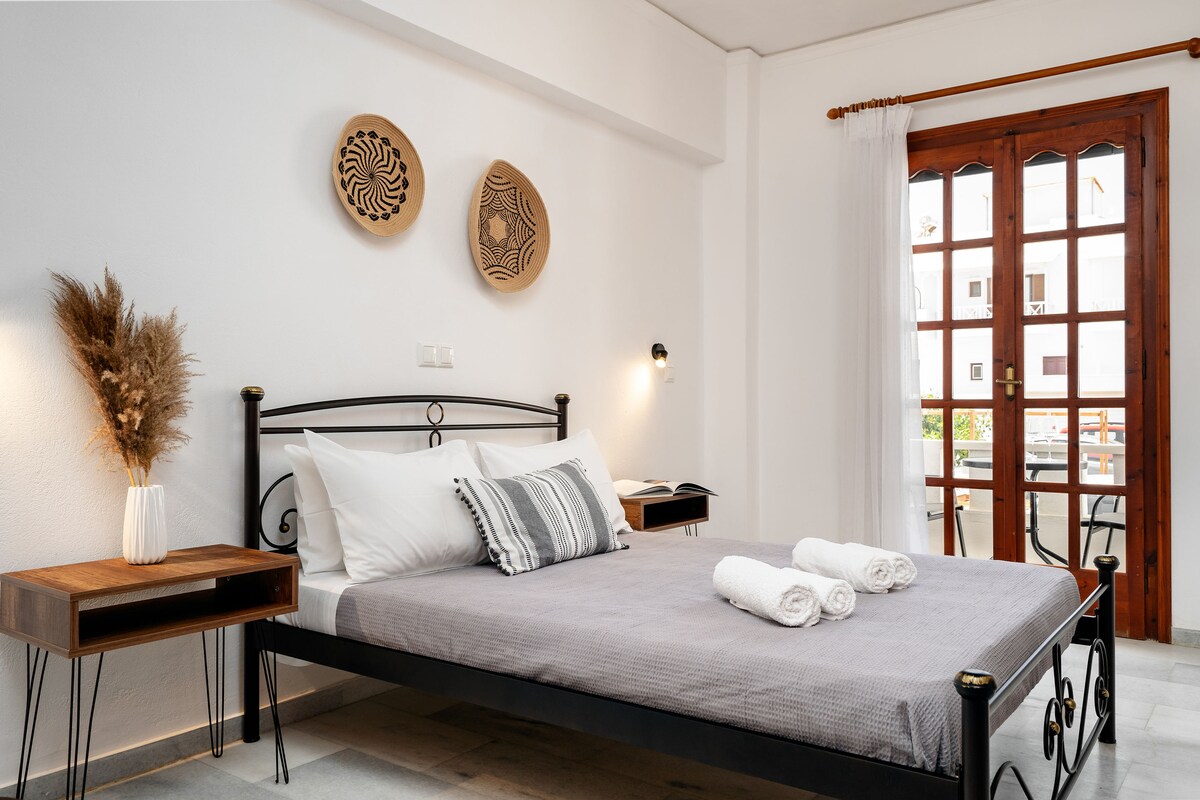 A bright and airy room features a spacious queen bed adorned with fresh linens. Two decorative wall pieces are mounted above the bed. Natural light enters through the wooden window, which leads to a patio, while a cozy bedside table and soft lighting provide additional charm.