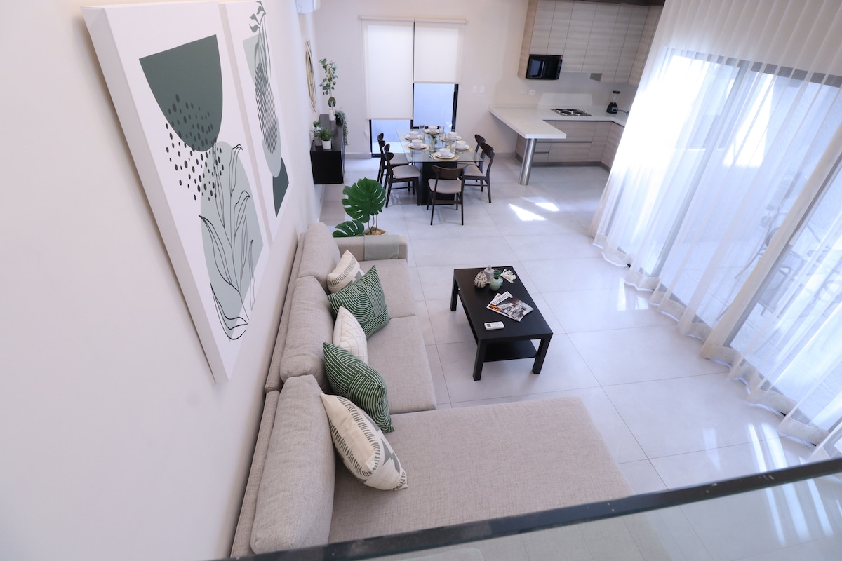 A modern living area features a light-colored sectional sofa adorned with green decorative cushions. A black coffee table sits in front, with magazines on it. The space is enhanced by large windows, allowing natural light to brighten the room and showcasing a dining setup in the background.