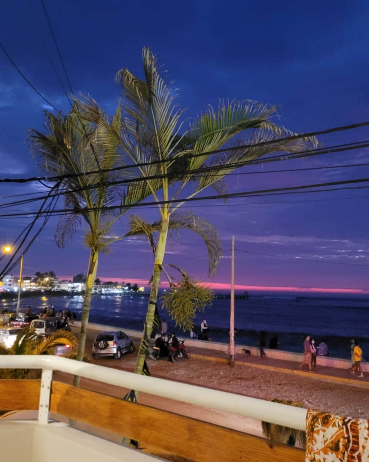 The view from the terrace features palm trees framing the scene against a twilight sky. The horizon displays a gradient of purple and pink hues, while the beach is visible below, with people walking and vehicles parked along the shore.