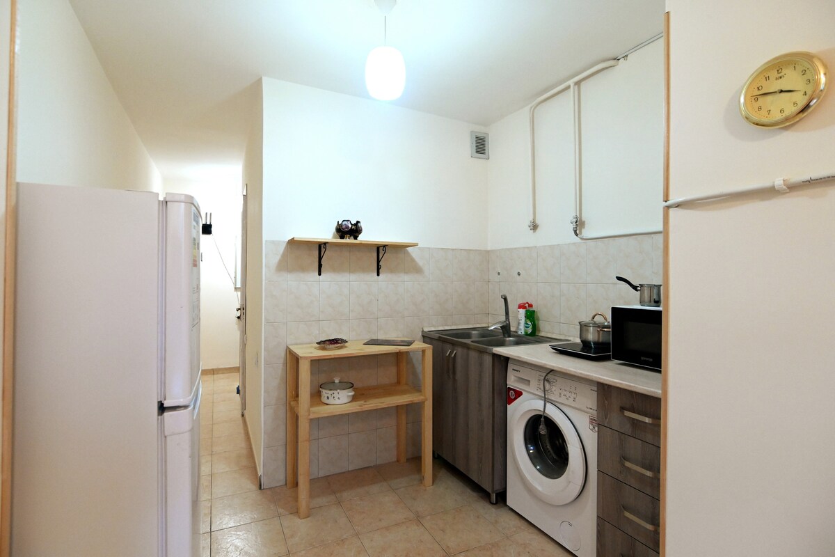 A functional kitchen features light-colored cabinetry and a stainless steel sink. Essential appliances, including a washing machine and microwave, are visible. A wooden table provides additional space, while a clock hangs on the wall, adding a practical touch to the modern design.