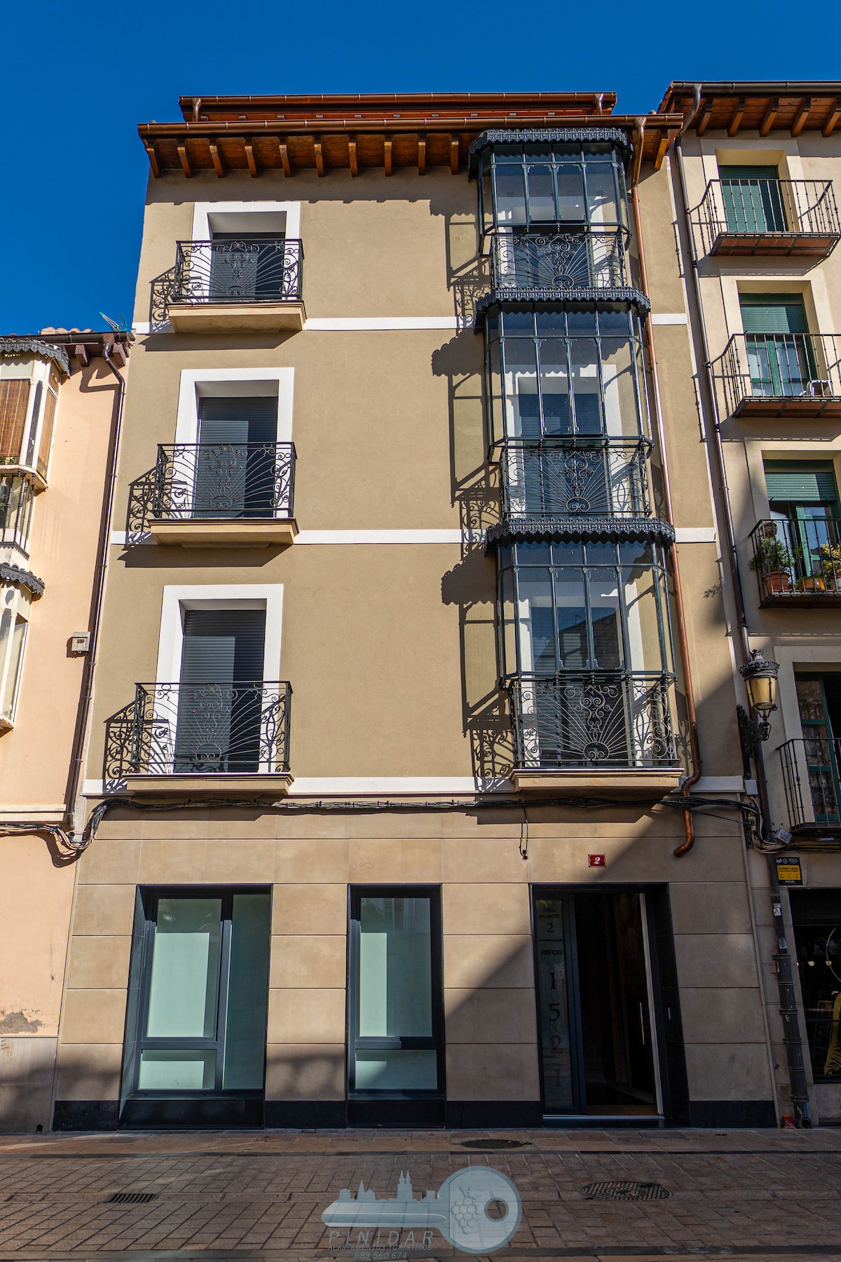 The building features a modern facade with three prominent balconies adorned with decorative metal railings. Large windows provide ample natural light, and the entrance is marked by a well-defined doorway. The ground floor showcases a clean, minimalist design.
