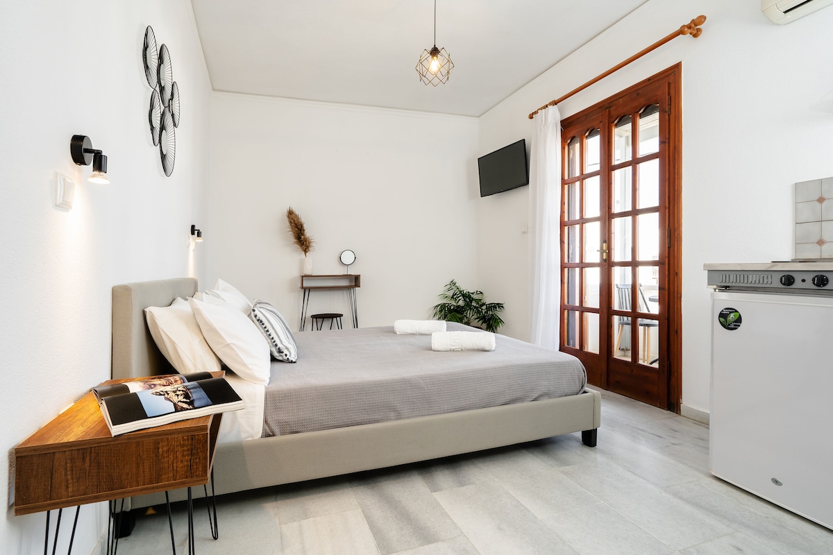 A spacious studio features a queen bed with fresh linens, complemented by a wooden side table. A kitchenette with a refrigerator is present, alongside a small dining area. Natural light enters through a set of double doors, which open to a balcony, enhancing the inviting setting.