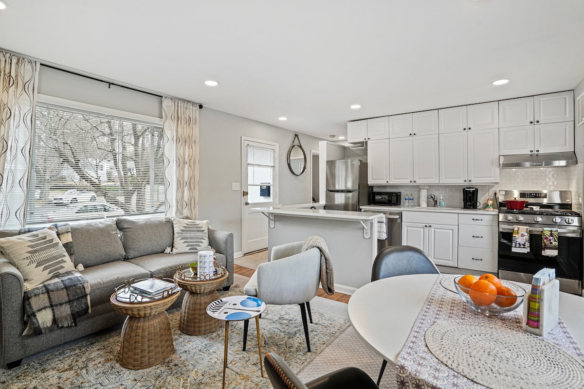 The living area features a comfortable grey sofa complemented by a patterned blanket. A round dining table is set with fruit, surrounded by modern chairs. The kitchen is equipped with new appliances, and ample natural light enters through large windows adorned with soft curtains.