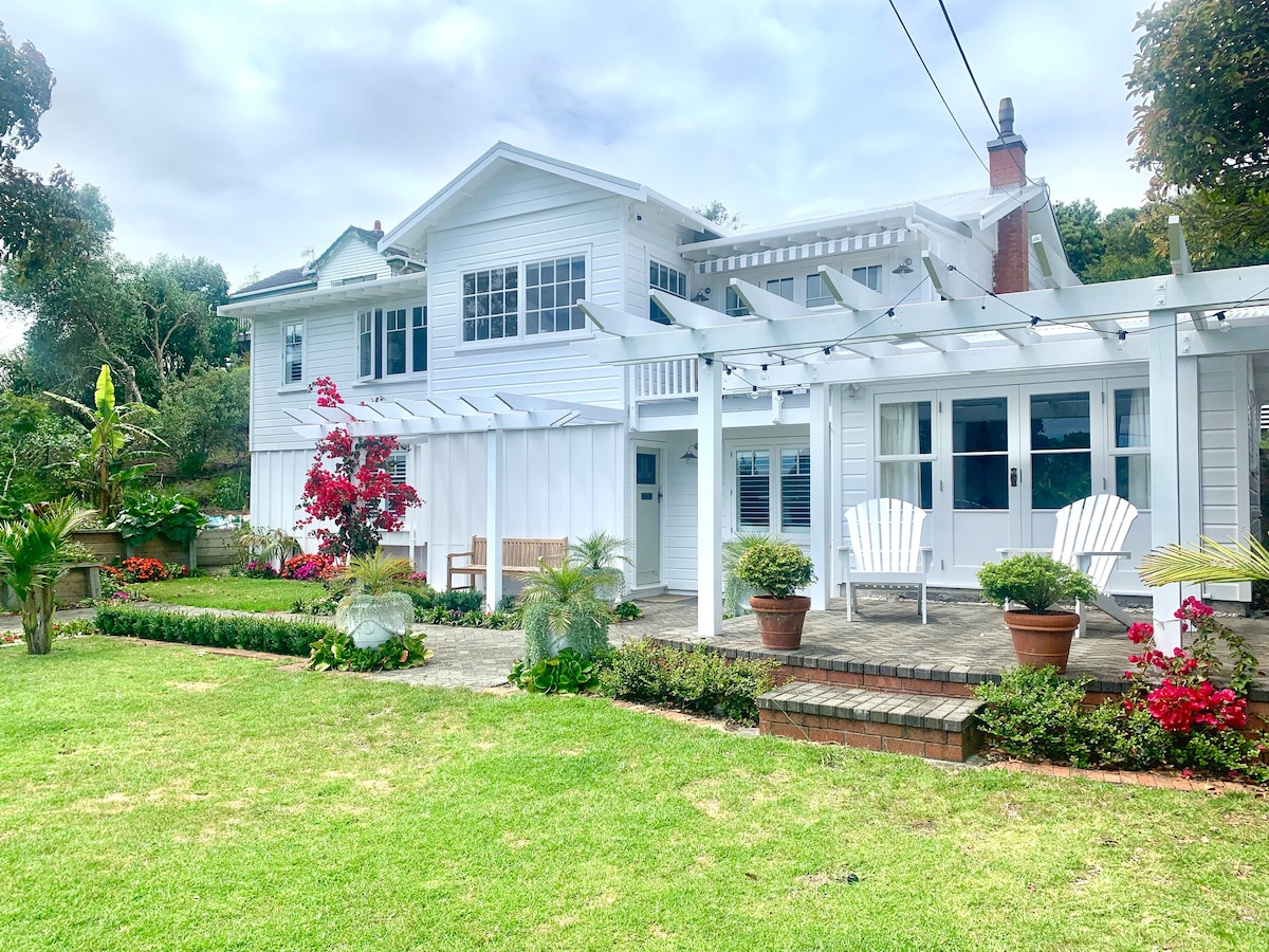 A charming exterior view of a two-story white house features a spacious patio with two Adirondack chairs. Lush green lawn surrounds the property, complemented by colorful flowerbeds and tropical plants. The structure showcases a pergola, adding architectural interest to the inviting outdoor space.