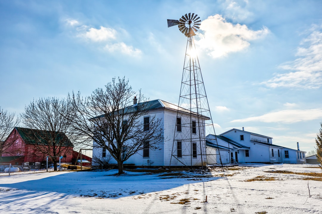 A white farmhouse sits amidst a snow-covered landscape, framed by leafless trees. A metal windmill stands tall, casting a shadow in the sunlight. Nearby, additional farm buildings are visible against a clear blue sky, creating a serene rural setting.