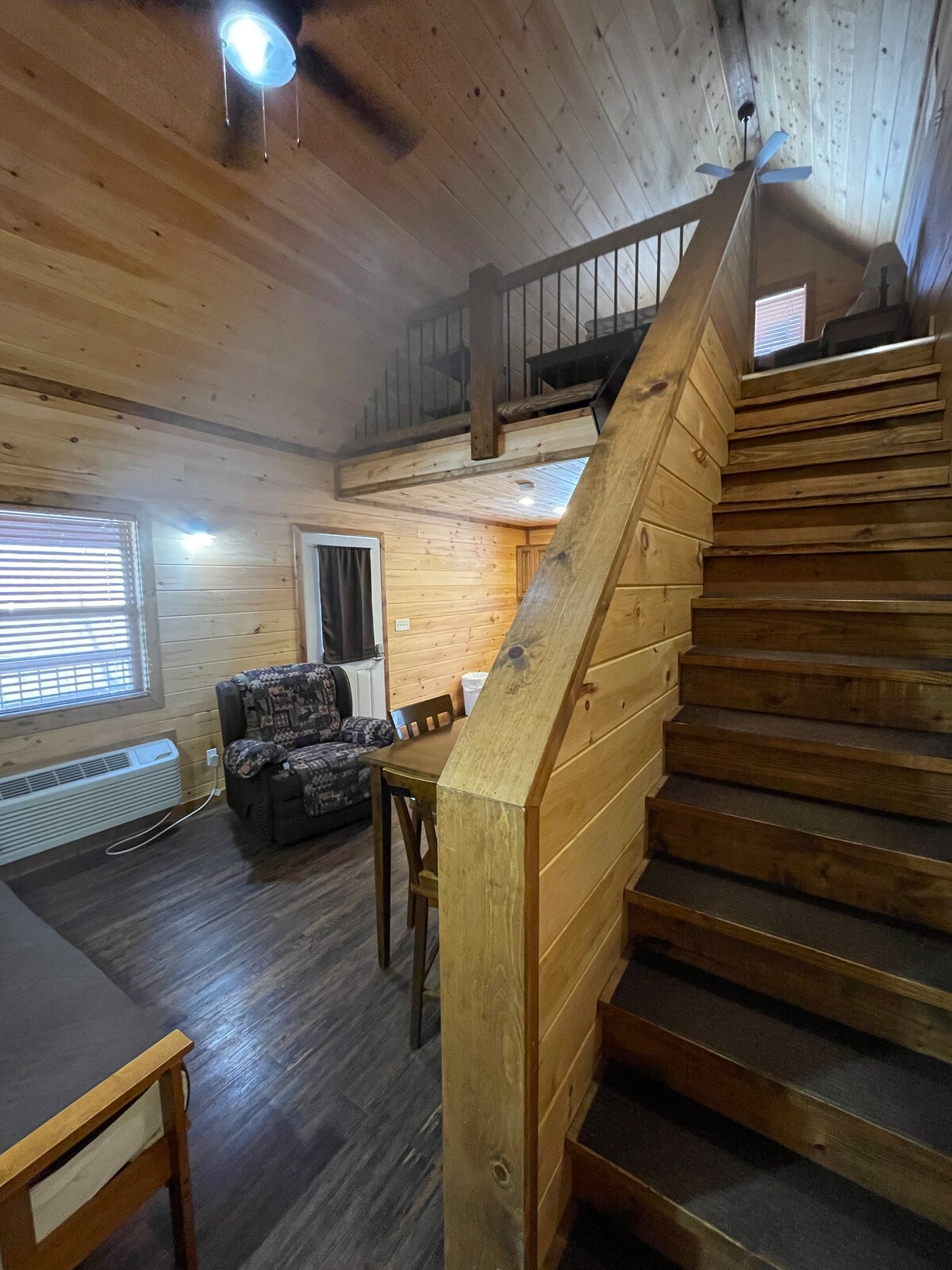 A wooden staircase leads to an upper loft area in a cozy cabin, illuminated by natural light. The main floor features a seating area with a patterned armchair and a small dining table. Wooden paneling decorates the walls, enhancing the rustic atmosphere.
