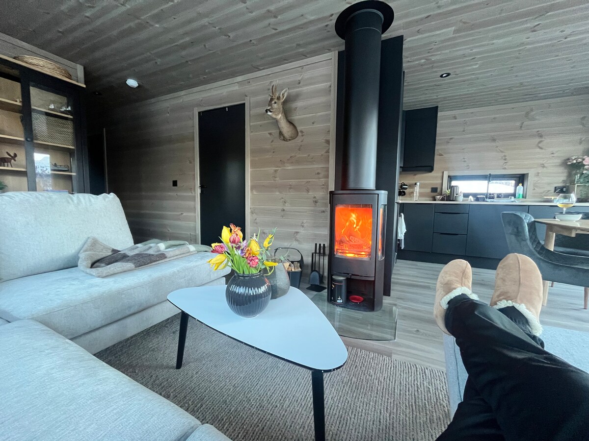 A cozy living area features a plush sofa complemented by a low coffee table. A decorative vase filled with flowers is placed on the table. A modern wood stove stands prominently, enhancing the warmth of the space. Natural light filters through the room.