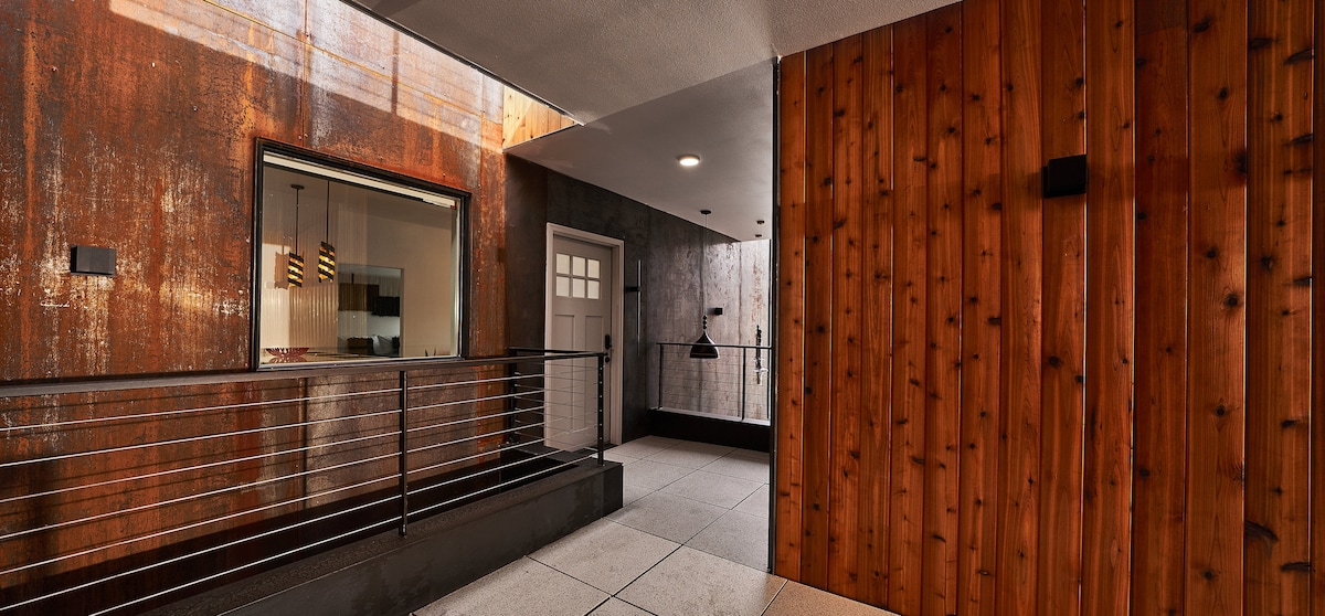 The entrance area features a combination of corten steel and cedar paneling, showcasing a warm wood tone against an industrial backdrop. A large window allows natural light to enter the space, while sleek metal railings provide safety along the walkway.