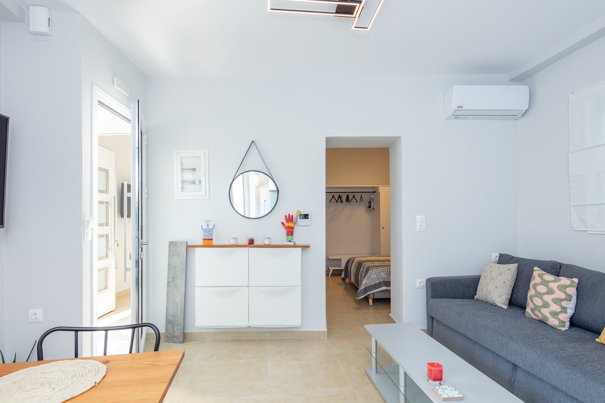 A modern living area features a dark gray sofa adorned with patterned cushions, complemented by a wooden dining table. A sleek mirror is mounted on the wall, and light enters through a nearby door. A hallway leads to a well-organized bedroom with hanging space.