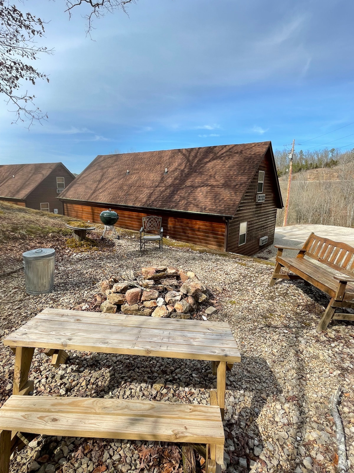 An outdoor area features a fire pit surrounded by seating on gravel, with a wooden picnic table and a bench in view. The cabin exterior is visible, with a sloped roof and an inviting arrangement of trees in the background.