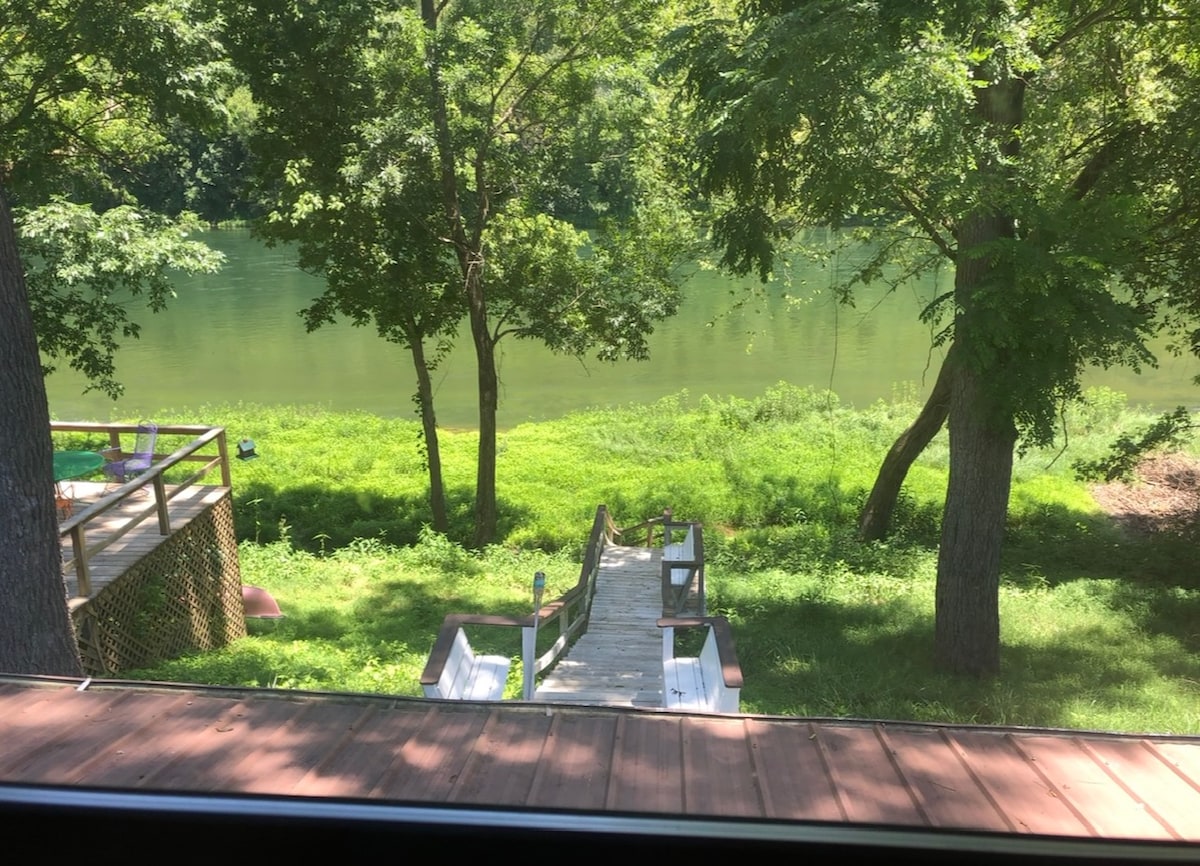 A view of the river can be seen from the cabin, framed by lush greenery and trees. A wooden staircase descends towards the water, surrounded by well-kept grass. A patio area is visible to the left, providing a peaceful setting by the shore.