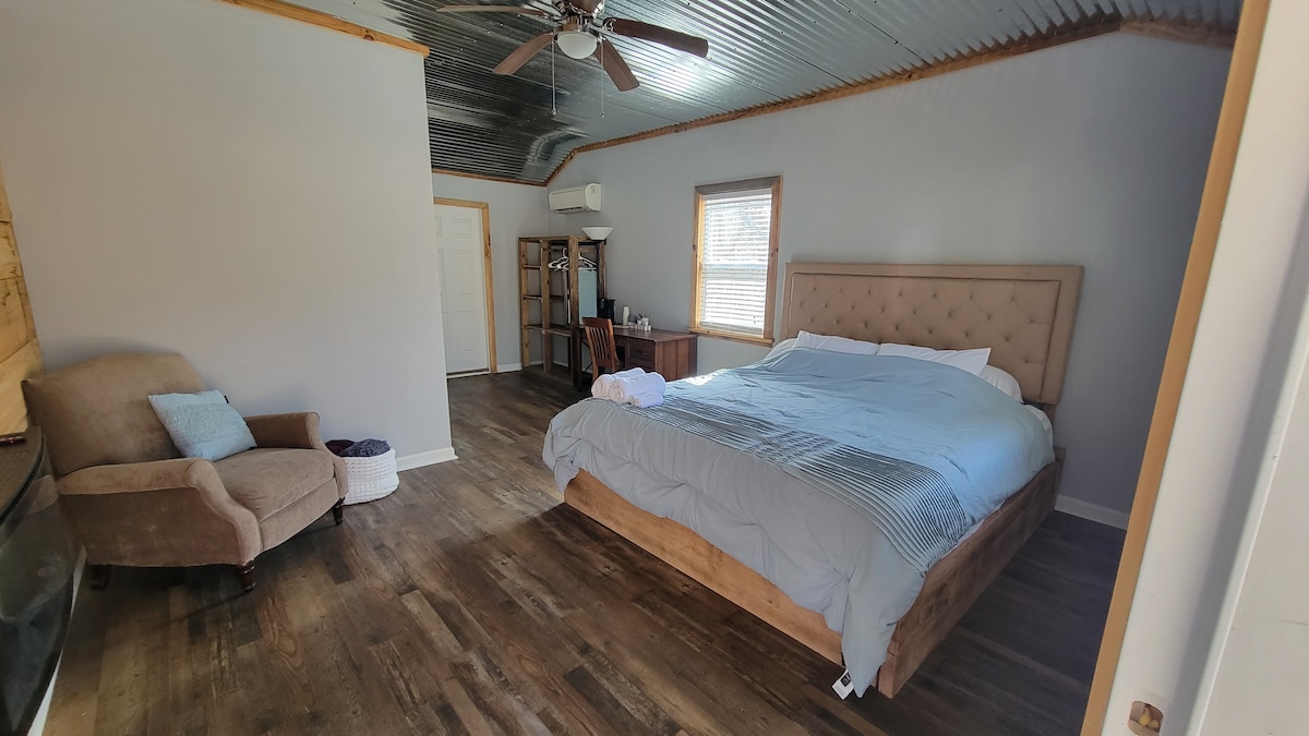 A spacious sleeping area features a king size bed with a tufted headboard and soft bedding. A comfortable armchair is positioned nearby, alongside a wooden desk and chair. Natural light flows in through a window, illuminating the wood flooring.