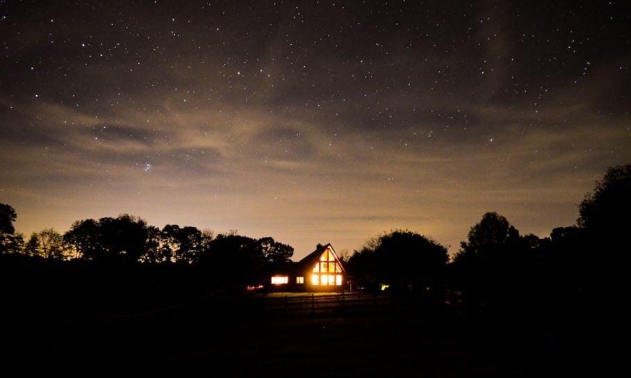 A charming house is illuminated against a night sky, surrounded by trees. The distant stars create a serene atmosphere, enhancing the peacefulness of the seven-acre property.