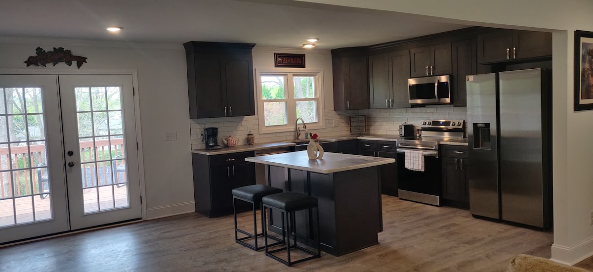A modern kitchen features dark cabinetry and stainless steel appliances, including an oven and refrigerator. An island with seating for two is centered in the room. Natural light enters through two windows and a glass door leading to a deck, enhancing the open feel.