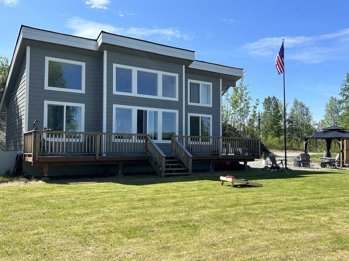 The modern chalet exterior is showcased with large windows framing the entrance. A spacious deck extends from the house, lined with wooden railings. A green lawn leads to the firepit area, while a flagpole displays the American flag, set against a bright blue sky.