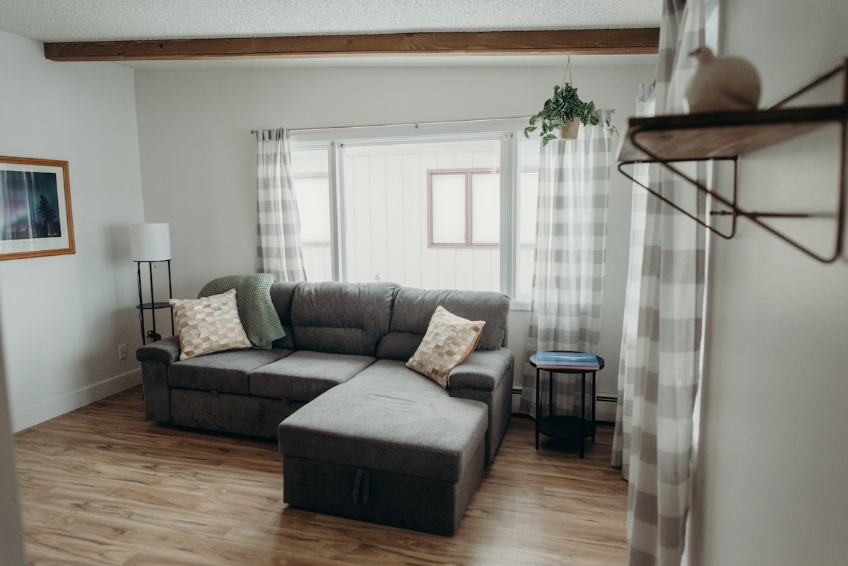 A comfortable living area is showcased with a sectional sofa and decorative pillows. Natural light filters through sheer curtains, illuminating the space. A small side table and a framed artwork add subtle charm to the room, inviting relaxation.