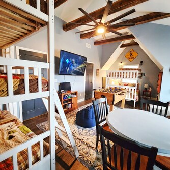 An inviting bunk/game room features two sets of bunk beds and a foosball table. A large screen TV is mounted on the wall, and a round dining table with four chairs is positioned nearby. Exposed wooden beams adorn the ceiling, enhancing the spacious feel.