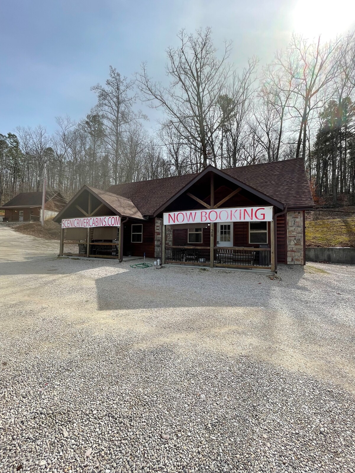 A charming cabin exterior is shown, featuring a wooden and stone facade under a sloped roof. Bright signage stating 'NOW BOOKING' is displayed prominently. A gravel driveway leads to the cabin, surrounded by a lightly wooded area with trees in the background.