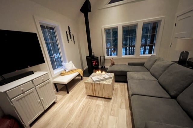 A cozy living area features a large sectional sofa and a small armchair, centered around a rustic coffee table. A fireplace stands nearby, contributing warmth. Large windows provide views of the surroundings, and a television is mounted on the wall across from the seating.