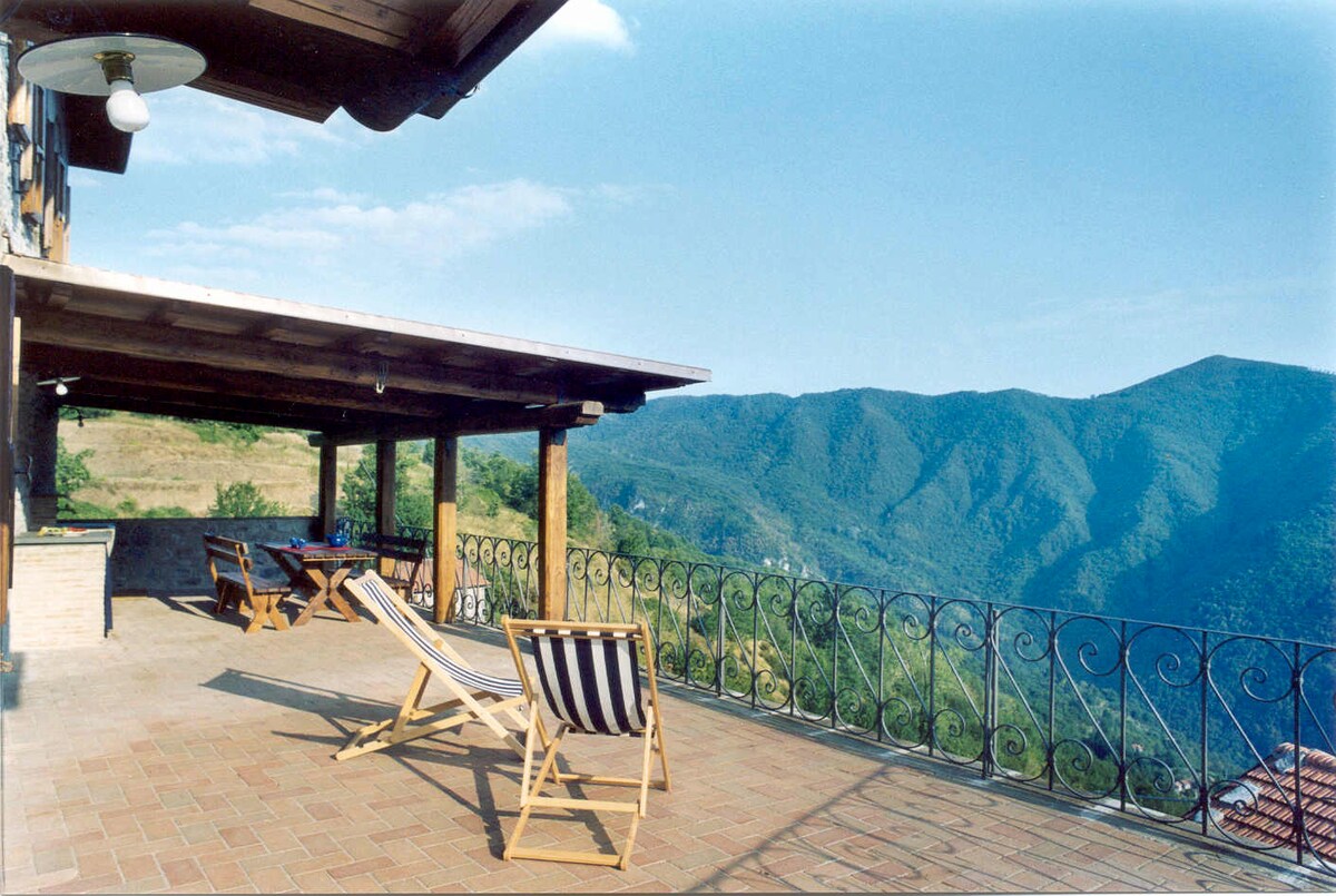 A spacious terrace is furnished with wooden lounge chairs and a dining table, offering a panoramic view of the surrounding mountains. The covered area provides shade, while the railing adds a decorative touch. Clear skies enhance the serene ambiance of the outdoor space.