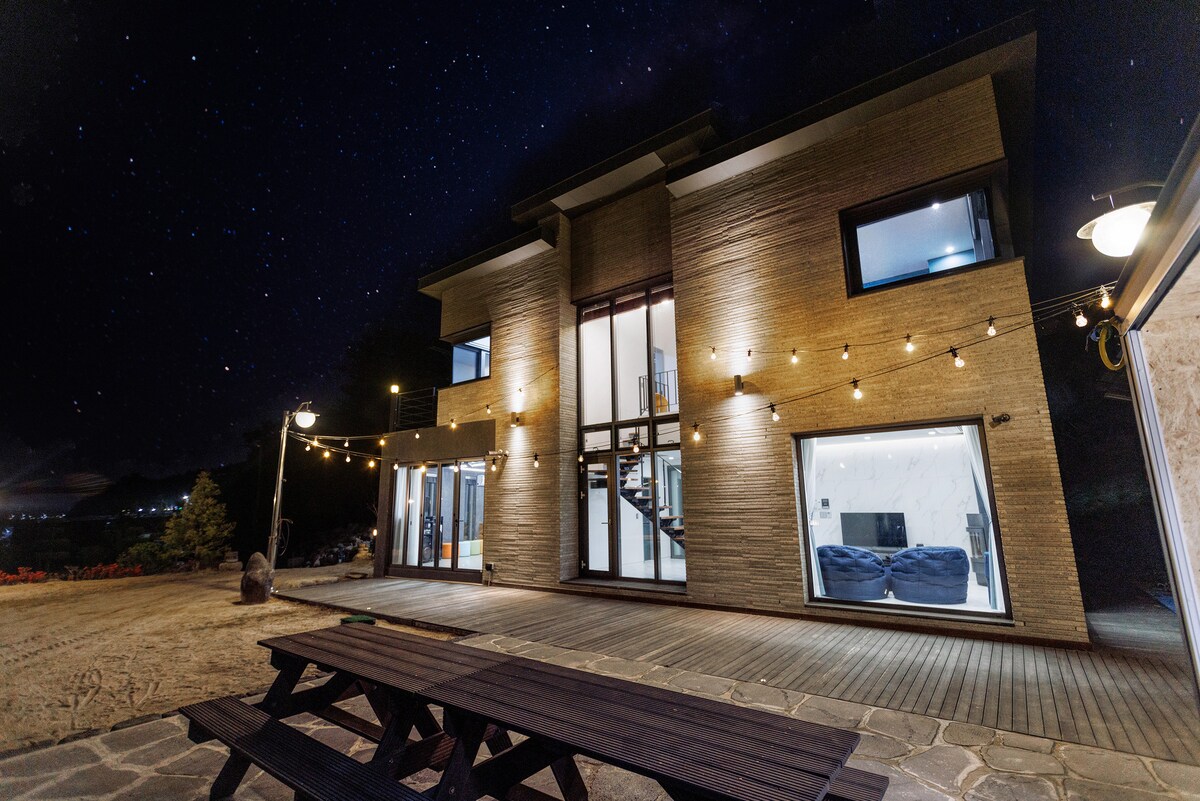 The two-story structure is illuminated against a clear night sky filled with stars. Large glass windows reflect the warm glow of outdoor lights, showcasing an inviting atmosphere. A wooden picnic table is situated in the foreground, enhancing the outdoor gathering space.
