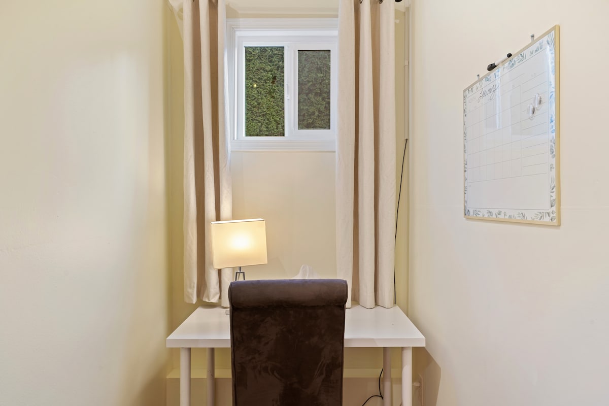 A dedicated workspace is presented with a simple white desk and a comfortable chair. A small desk lamp provides lighting, while light curtains frame a window that allows natural light to brighten the area. A whiteboard is mounted on the wall for notes or reminders.