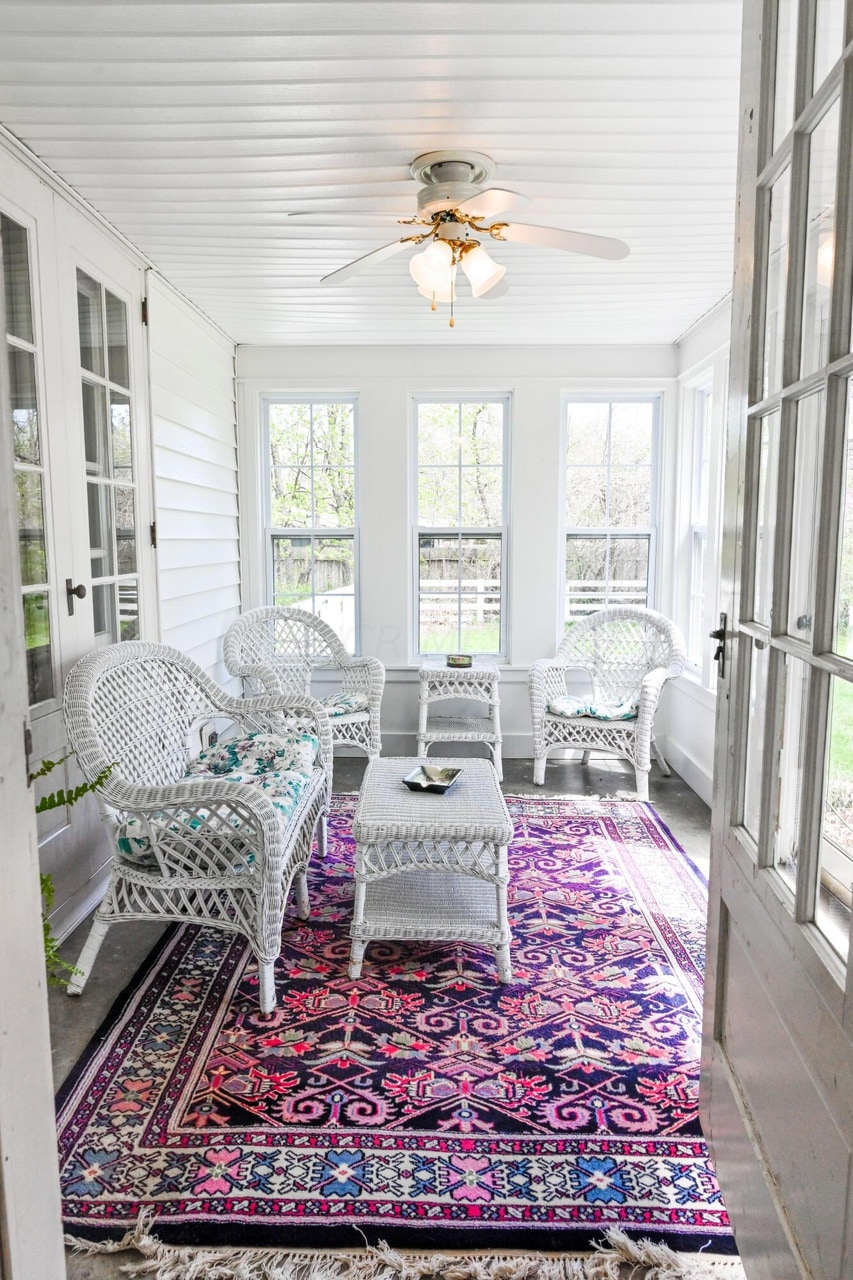 A sunlit porch features wicker furniture arranged on a colorful rug. Large windows surround the space, allowing plenty of natural light. A ceiling fan provides airflow. A small table sits in the center, enhancing the inviting atmosphere for relaxation.