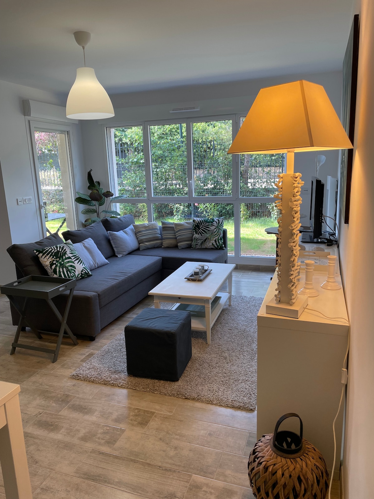 A living area features a spacious grey sofa adorned with decorative cushions, accompanied by a small black ottoman and a white coffee table. Natural light filters through large glass windows, offering views of the garden outside. A standing lamp and decorative elements provide additional ambiance.