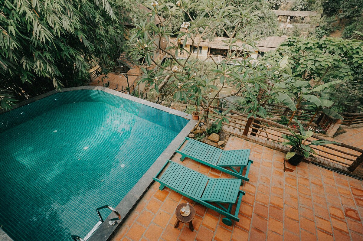 A serene outdoor area features a turquoise pool surrounded by lush greenery. Two wooden lounge chairs sit alongside the pool on a terracotta tiled deck, with an inviting table positioned nearby, creating a tranquil space for relaxation.