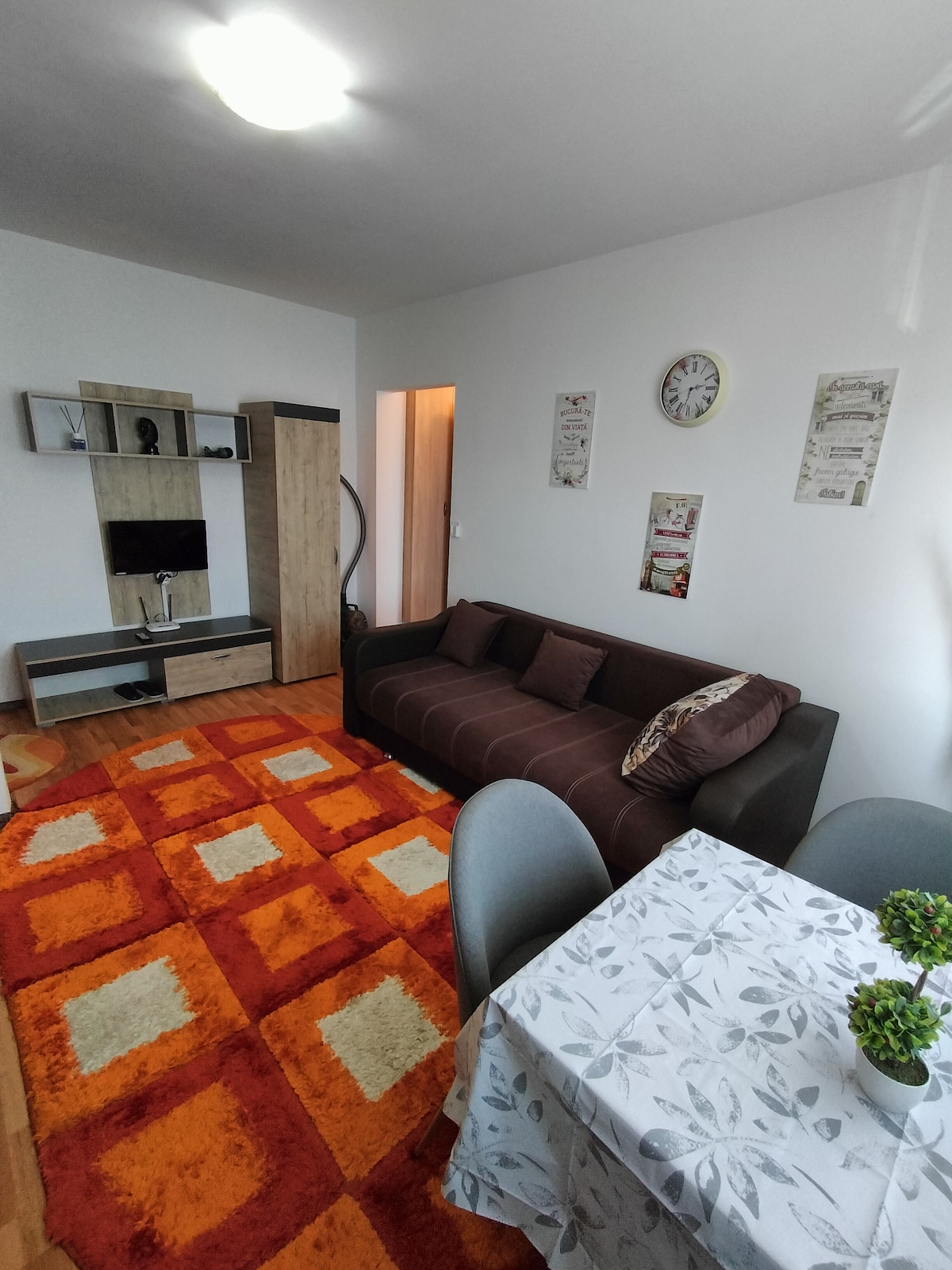 A cozy living space features a dark sofa on an orange and brown patterned rug. A small dining table with light-colored cloth sits beside it, accompanied by two chairs. A media console houses a television, and wall decor adds character to the bright room.
