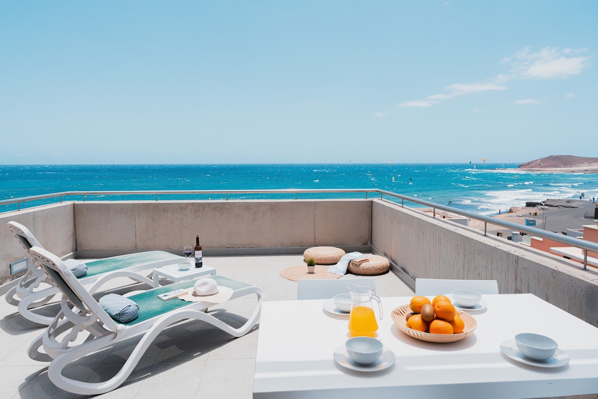 An expansive terrace overlooks a vibrant blue sea, offering panoramic views of the beach and windsurfing activities. A modern dining table is set up with orange juice and fresh fruit, while comfortable loungers are positioned for relaxation under the bright sky.