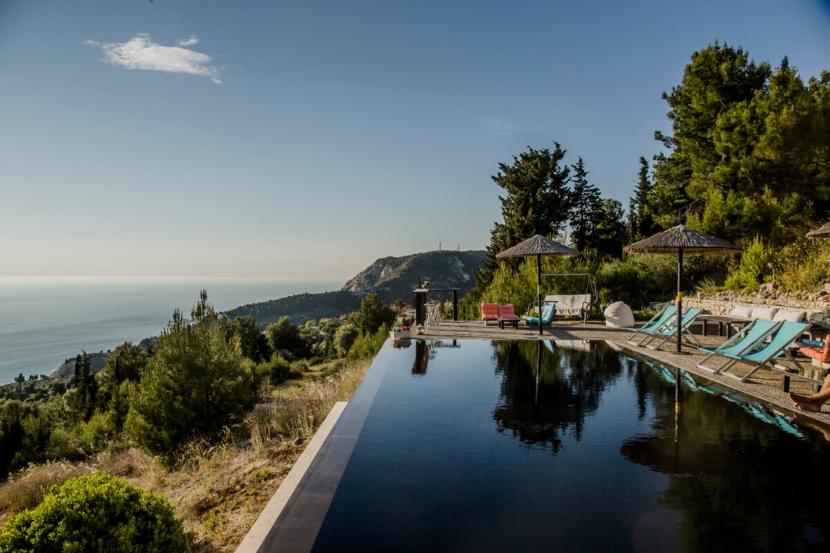 A serene infinity pool reflects the sky, with sun loungers positioned nearby. Lush greenery and natural landscapes frame the view, leading to distant mountains and the shimmering sea. Umbrellas provide shaded areas, enhancing the tranquil outdoor environment.