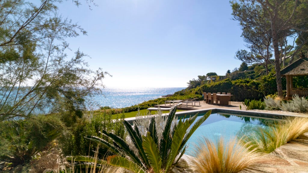 A tranquil outdoor area features a rectangular swimming pool surrounded by lush greenery. The shimmering Mediterranean Sea is visible in the background, creating a serene atmosphere. A wooden deck with seating options is positioned nearby, providing a space for relaxation.