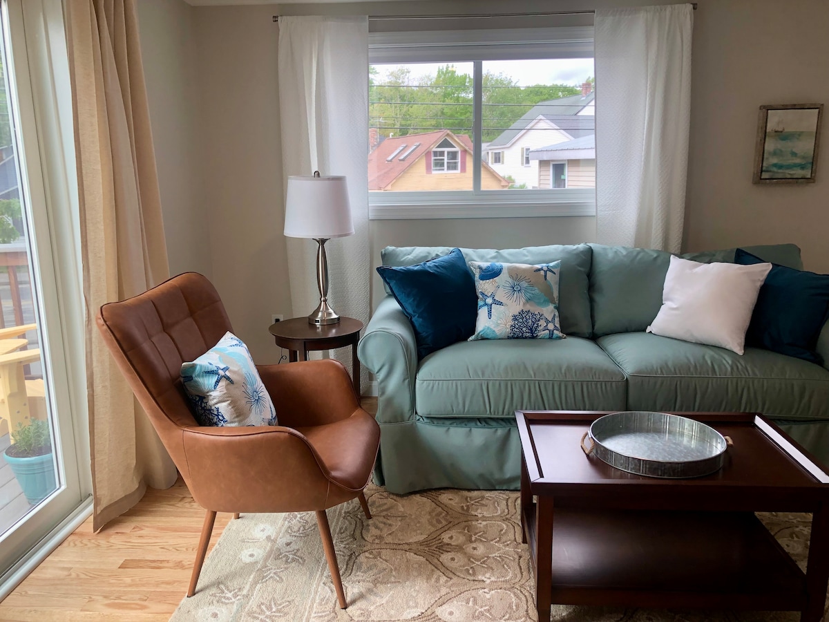 A comfortable living area features a soft green sofa accented with decorative pillows. A sleek brown chair complements the space. Natural light filters through a large window, adorned with light curtains, showcasing a view of the surroundings. A wooden coffee table sits in front.