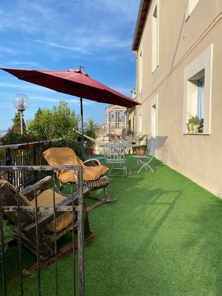 A shaded terrace is presented, featuring an umbrella and metal chairs surrounding a small table. A pair of comfortable rocking chairs add to the seating options. Lush green artificial grass carpets the space, enhancing the sense of tranquility against the backdrop of nature.